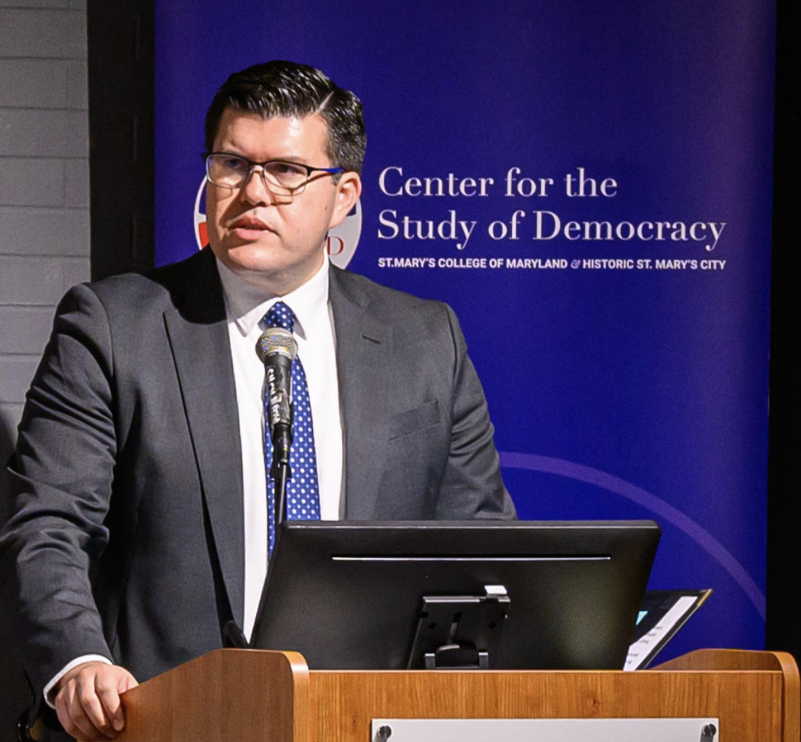 A man in a suit speaks at a podium with a microphone in front of a sign for the 民主研究中心 at St. Mary's College of Maryland.