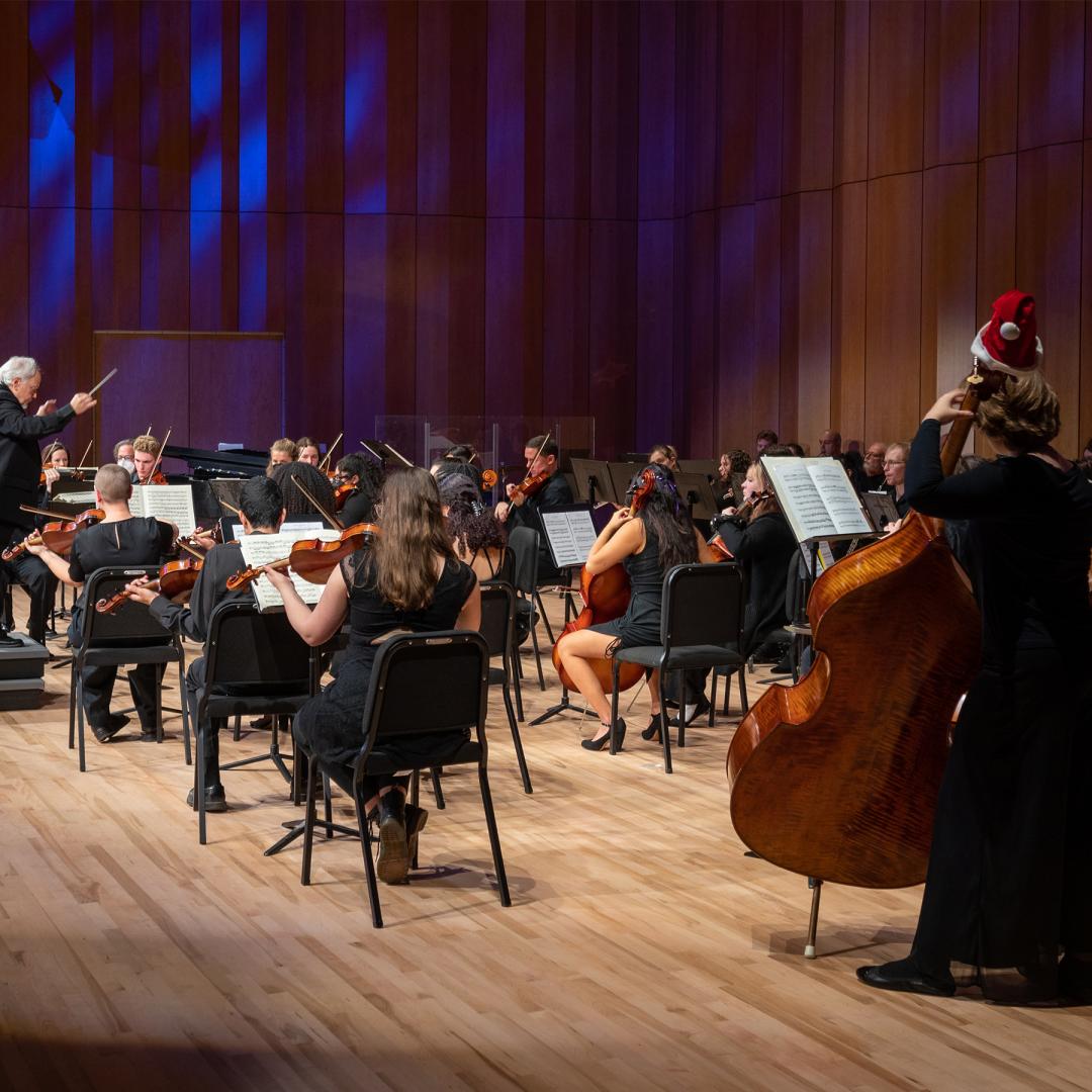 A symphony orchestra performs on stage with a conductor. One musician in the front right wears a Santa hat while playing a double bass.