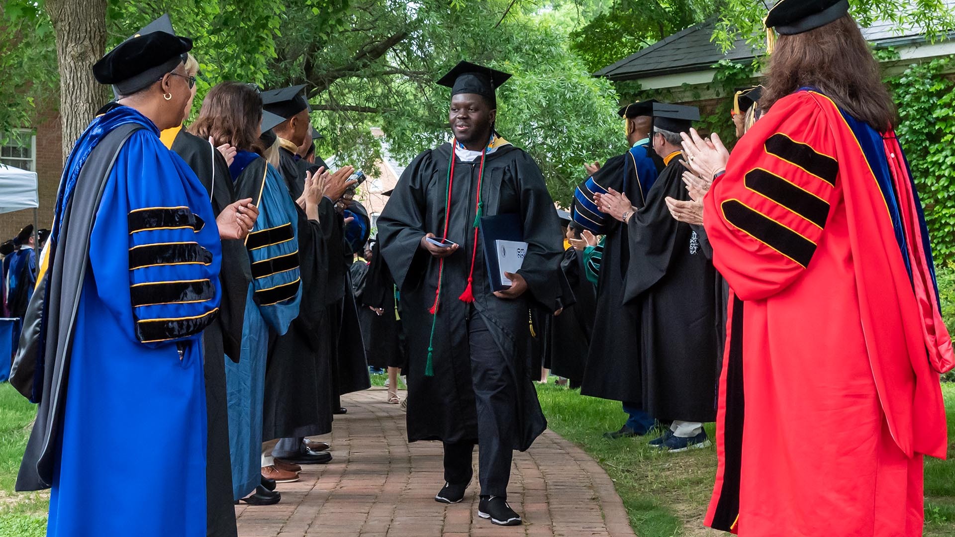 A graduate in cap and gown walks down a brick path lined with applauding faculty members in colorful academic regalia. The setting is outdoors with lush green trees and grass.