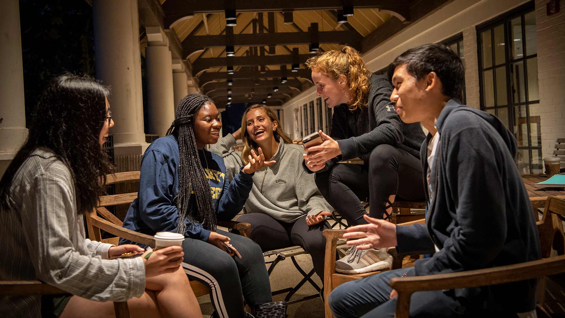 A group of five people sit in a circle on wooden chairs in an outdoor setting, engaged in lively conversation. They are smiling and appear to be enjoying each other's company. One person holds a coffee cup. The setting is warmly lit.