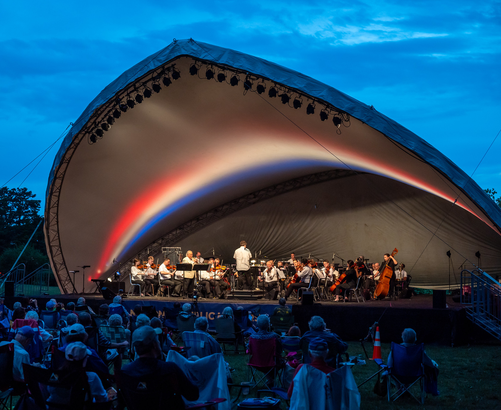 Orchestra performing on an outdoor stage under an illuminated, curved canopy during twilight. Audience members sit in chairs on the grass, while the sky transitions from blue to dusk.