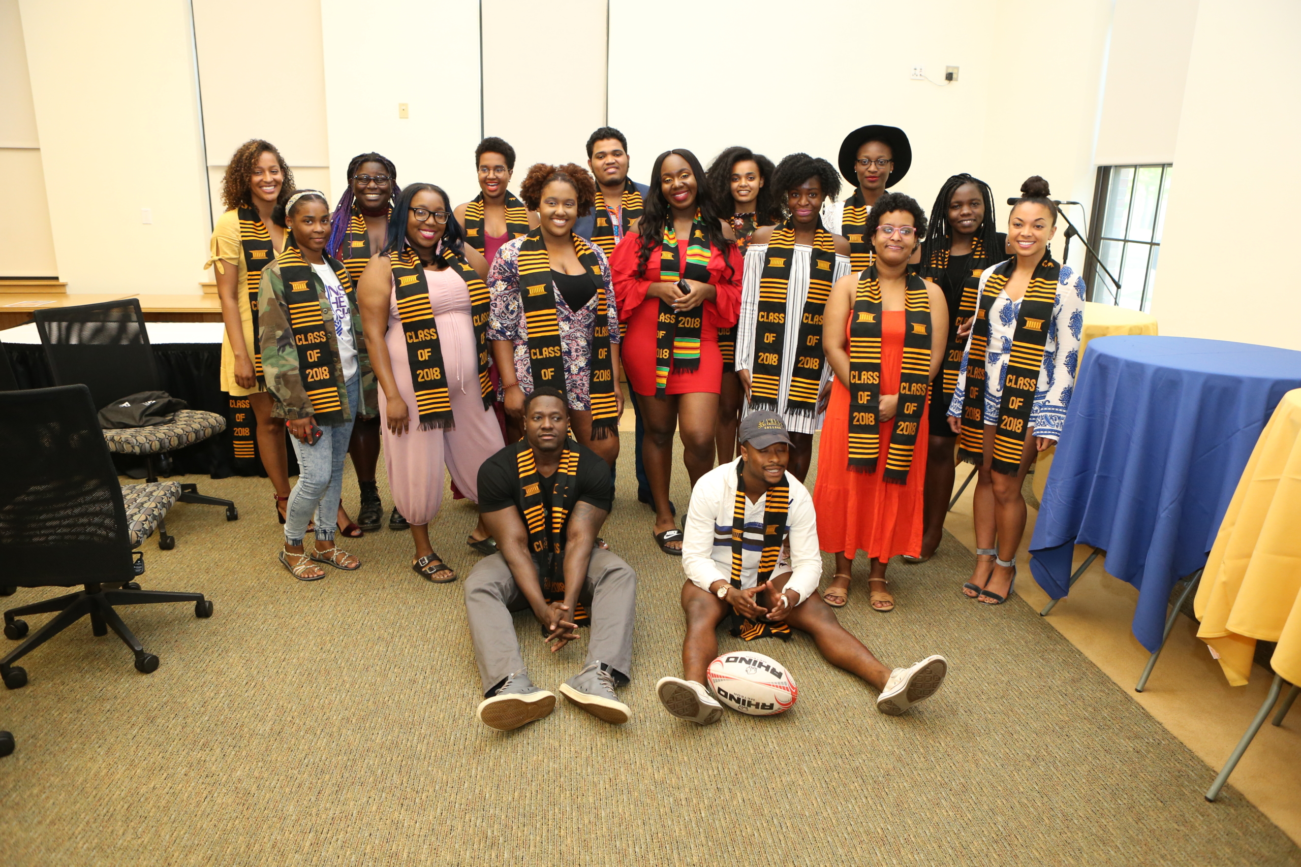 A group of nineteen people, mostly standing and some sitting on the floor, are dressed in a mix of formal and casual clothing. They are wearing vibrant sashes with colorful patterns and "Class of 2018" text. A rugby ball is on the floor in front.
