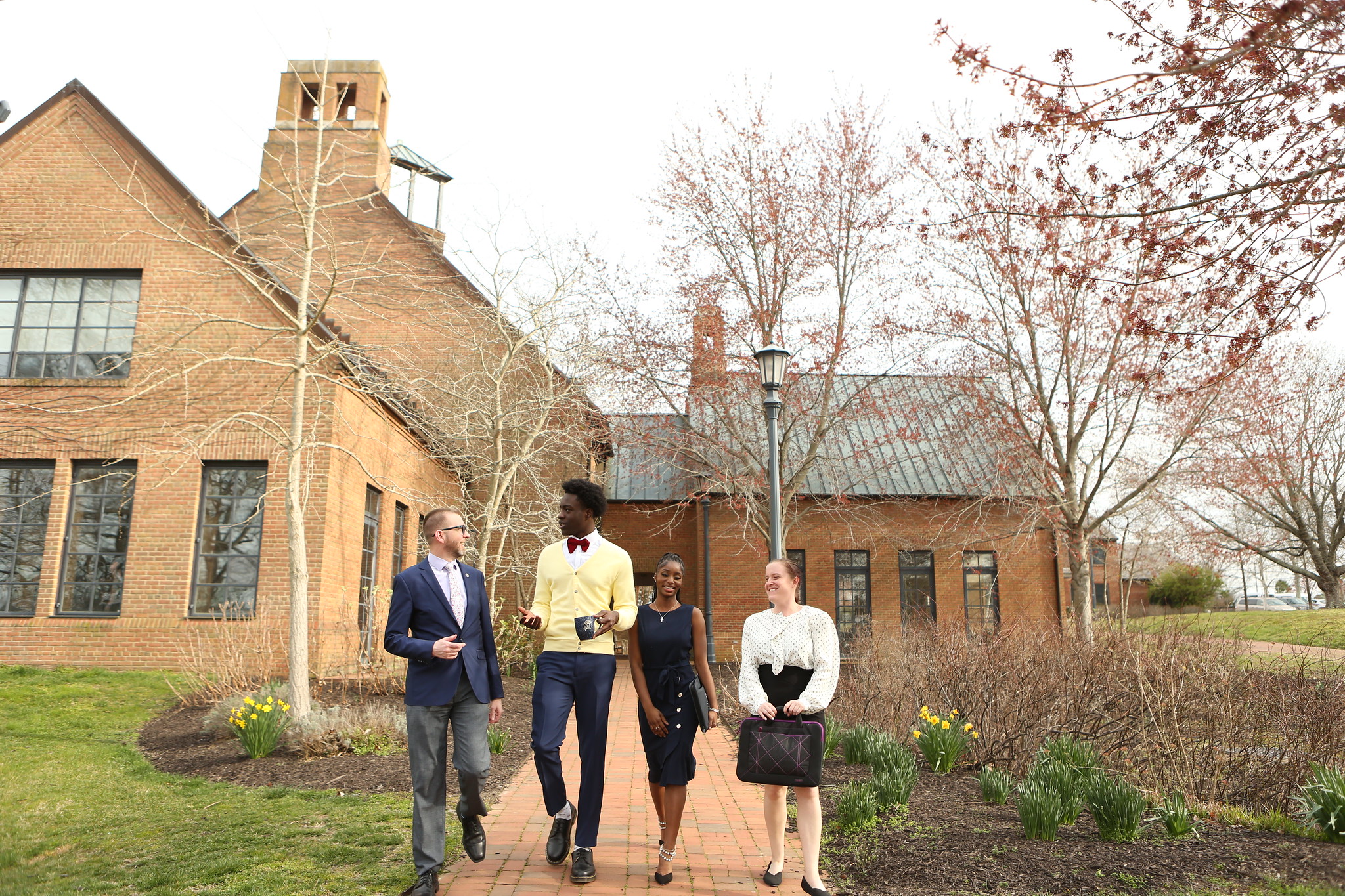 Four people, three wearing business attire and one in a casual sweater, walk on a brick path next to a brick building with a tall chimney. Leafless trees and daffodils are in the background. The group appears cheerful and engaged in conversation.
