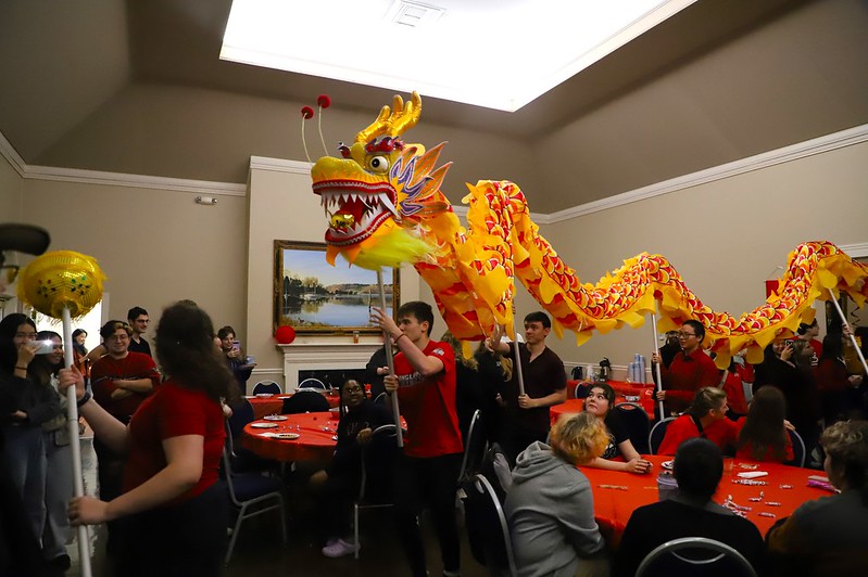 A vibrant dragon costume is paraded by a group of people inside a room. Several seated individuals watch the performance. The dragon is bright yellow with red patterns. The room has red tablecloths and a painted landscape hangs on the wall.