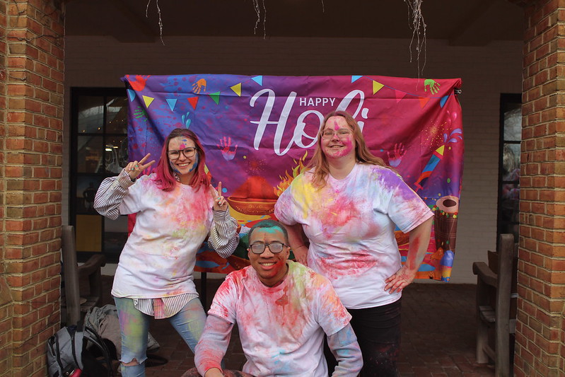 Three people covered in colorful powder smile in front of a "Happy Holi" banner. Two stand while one kneels. They are wearing white shirts splattered with vibrant colors, celebrating the Holi festival.