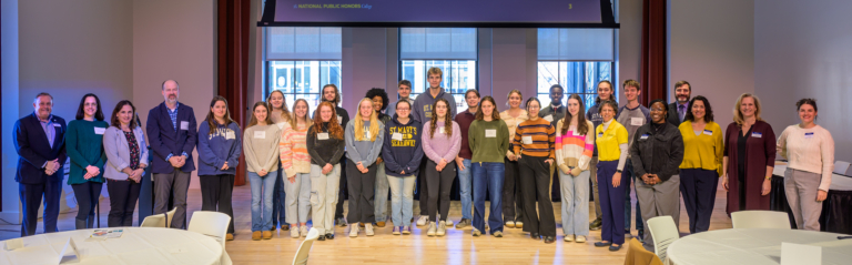 A diverse group of people, including adults and students, stand together in a well-lit room with large windows.