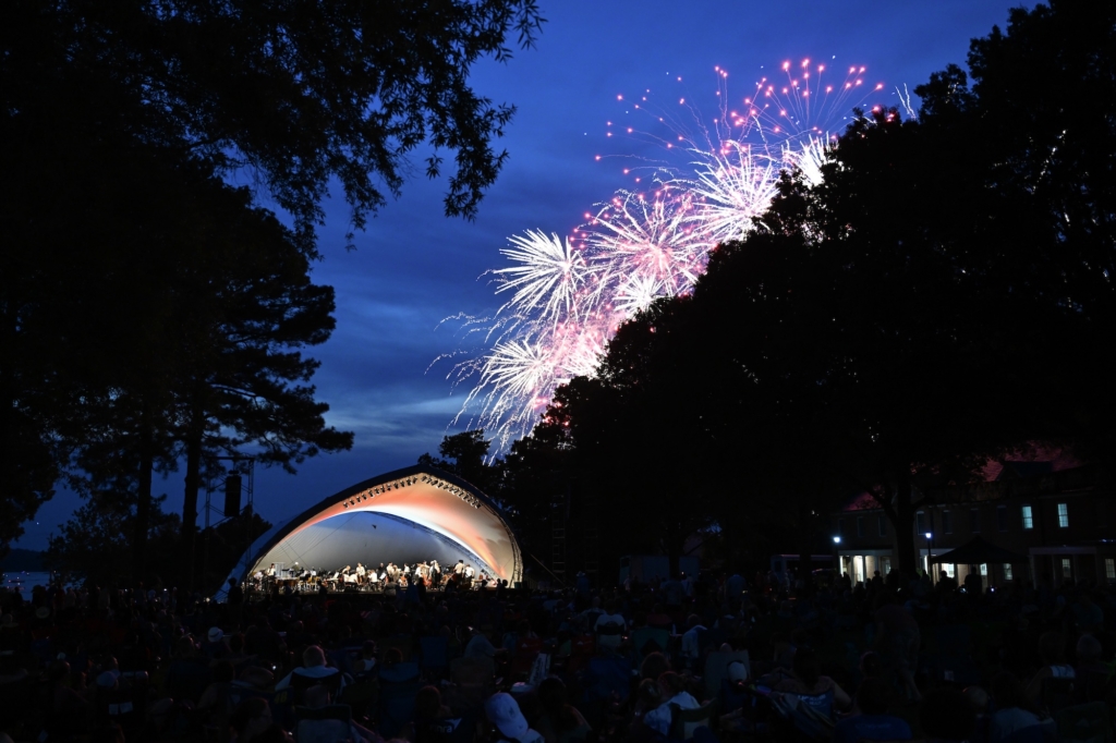 Fireworks light up the sky over an outdoor concert with a shell-shaped stage surrounded by trees and a crowd of people seated and watching.