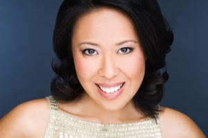 Woman with shoulder-length dark hair wearing a sleeveless, patterned beige top, smiling at the camera against a dark background.