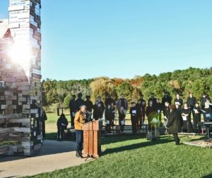 A woman speaks at a wooden podium in front of a tall sculpture composed of stacked wooden beams. A group of people observes nearby, seated and standing. The sun shines brightly, reflecting off the sculpture. Trees are visible in the background.