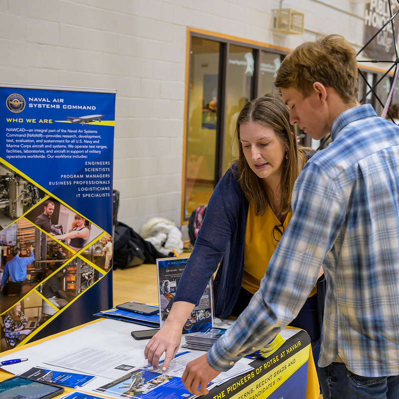 A woman explains information to a young man at a Navy recruitment booth. The booth has banners 和 brochures about Naval Air Systems Comm和. They are indoors, surrounded by tables 和 other booths.
