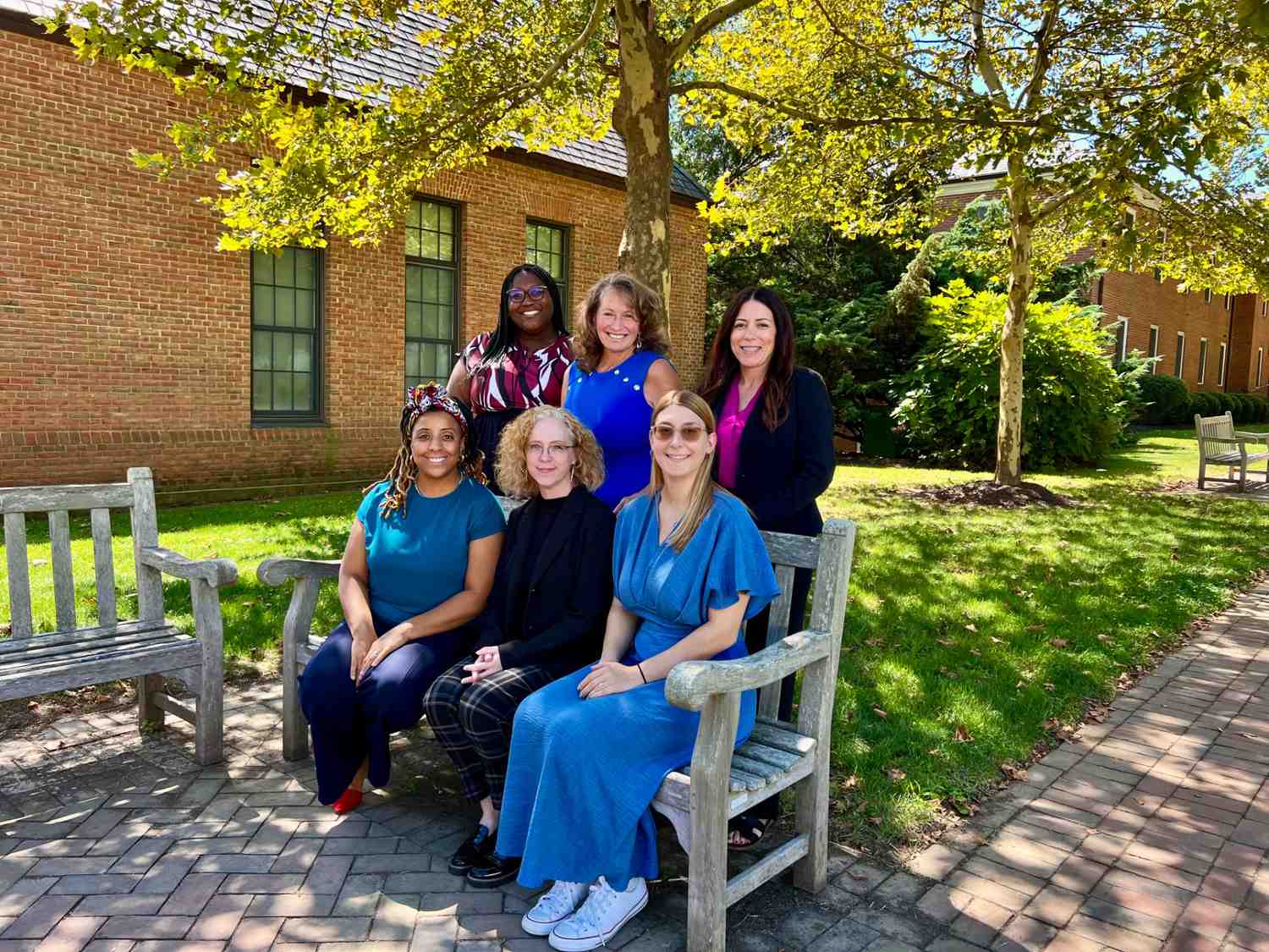 Six women pose for a group photo on 和 around a wooden bench outside, with trees 和 brick buildings in the background on a sunny day.