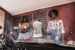 Three people grill food outdoors near a brick wall with bicycles in the background."