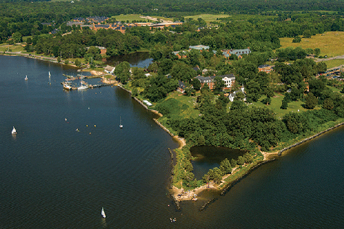 Aerial view of a lush, green shoreline with several houses surrounded by trees. Small sailboats are scattered across the water, and a dock extends into the bay. A mix of open fields and forested areas can be seen in the background.