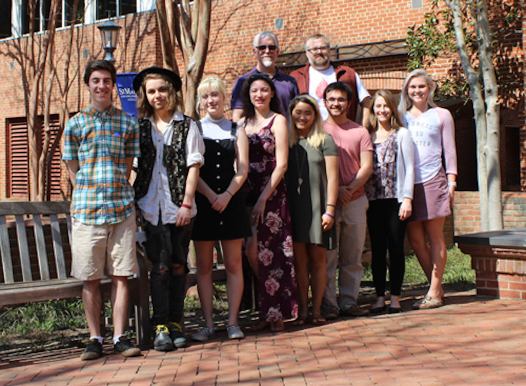A group of ten people standing outdoors on a brick path in front of a brick building and trees. They are smiling, and some are wearing casual clothing while others wear dresses or vests. The weather appears sunny.