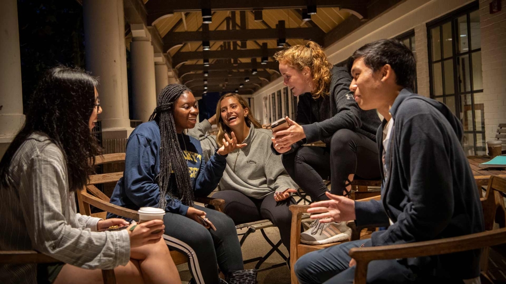 Five people are sitting in a circle outdoors on wooden chairs, engaged in lively conversation. They appear relaxed and happy, with one holding a phone and another holding a coffee cup, set against a background of columns and wooden beams.