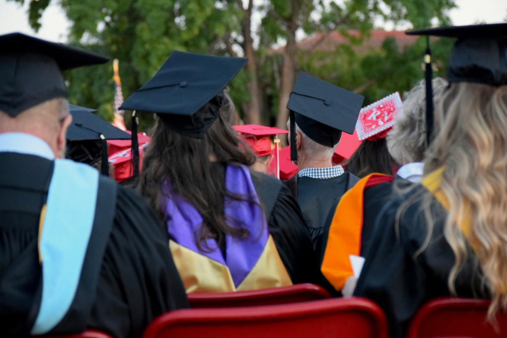 A group of graduates in black gowns and caps sit outdoors in red chairs. Some wear colored sashes and hoods; one has a decorated cap. Trees are visible in the background.