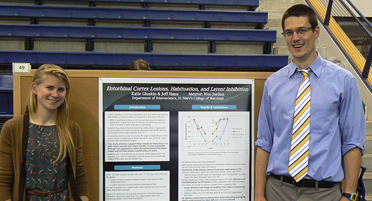 Two people stand next to a science poster titled "Entorhinal Cortex Lesions, Habituation, and Latent Inhibition" in a gymnasium setting. The woman on the left wears a patterned dress, and the man on the right wears a light blue shirt and tie.