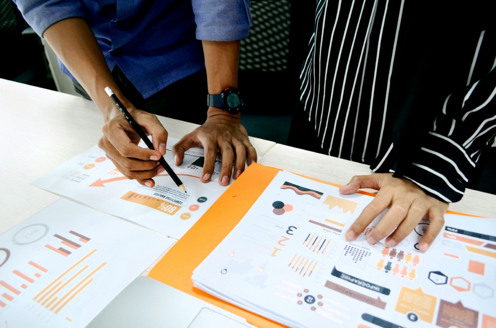 Two people are collaborating over documents and charts on a table. One person is writing with a pencil, while the other holds papers with graphs. Their clothing includes a blue shirt and a black-and-white striped top.