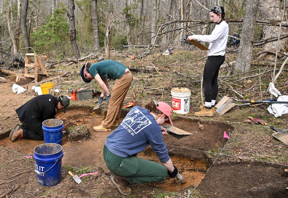 Four people digging in the ground outside while one person stands nearby holding a clipboard, observing the activity.