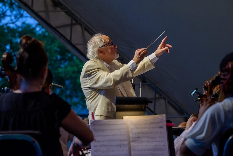 A conductor in a light-colored suit leads an orchestra during an outdoor performance. The musicians are seated with sheet music visible, while the conductor uses a baton, and trees are glimpsed in the background evening light.