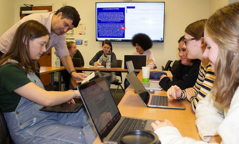 A group of students seated at tables work on laptops in a classroom. An instructor leans over to assist one student. A presentation screen displays text. Everyone appears engaged in the learning activity.