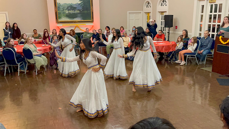 A group of women in white traditional dresses performs a dance in a community hall. They are surrounded by an audience seated at round tables decorated with red tablecloths. A speaker stands at a podium in the background.