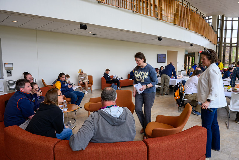 People are gathered in a spacious, modern lounge area. Some are seated on orange couches, engaged in conversation, while others are standing. Large windows and a wooden railing are visible in the background. Casual atmosphere with diverse activities.