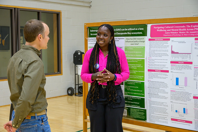 A woman presents research in front of a poster board. She is smiling and speaking to a man, who is listening attentively. The background shows a gymnasium setting. The posters contain graphs and text related to scientific research.