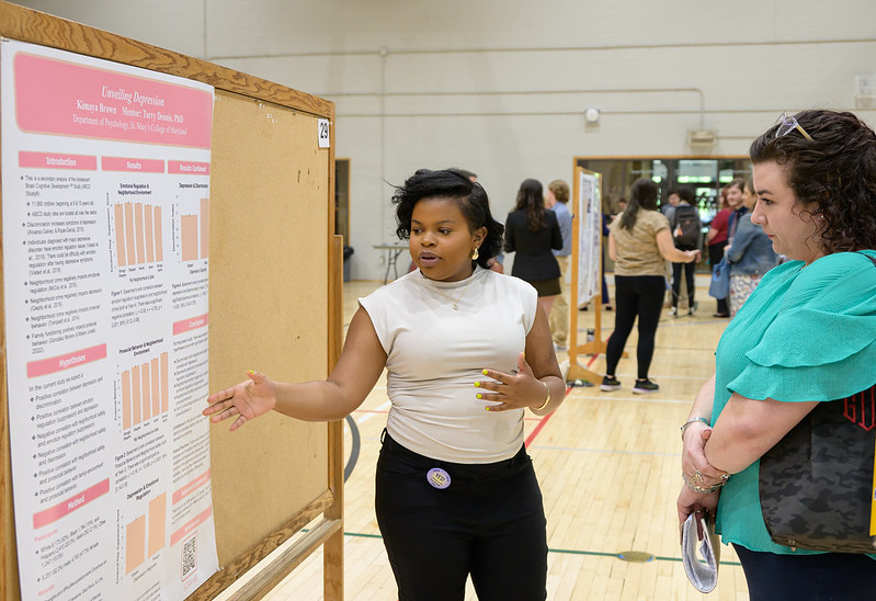 A woman stands beside a presentation board with graphs, explaining something to another woman who is listening attentively. They are in a gymnasium setting with other people and boards in the background.