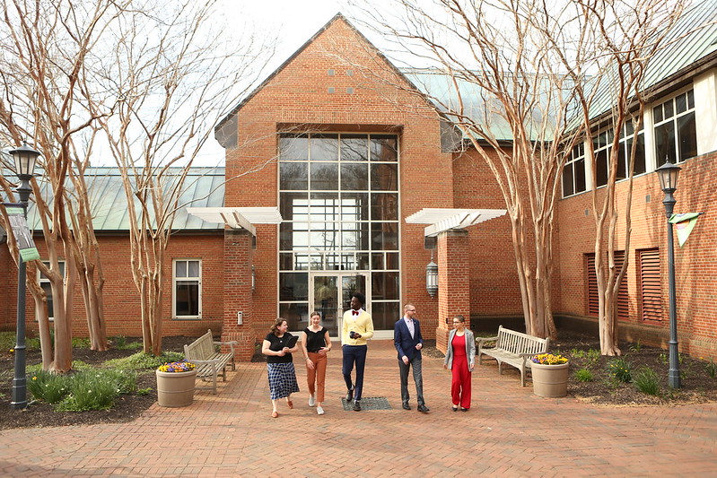 Five people walk together outside a brick building with large windows, surrounded by leafless trees, benches, and planters with flowers.
