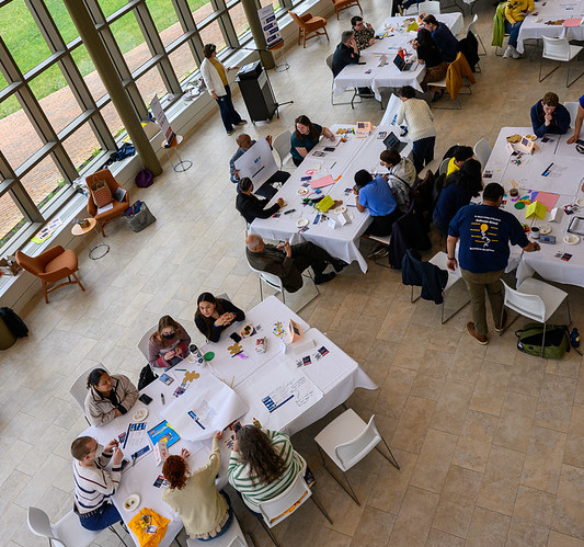 People sit at round tables in a large, bright room with floor-to-ceiling windows, engaging in group activities or discussions.