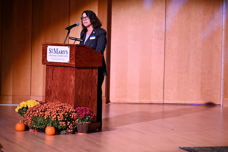 A woman stands at a podium labeled "St. Mary's College of Maryland," speaking into a microphone on a stage decorated with flowers and pumpkins.