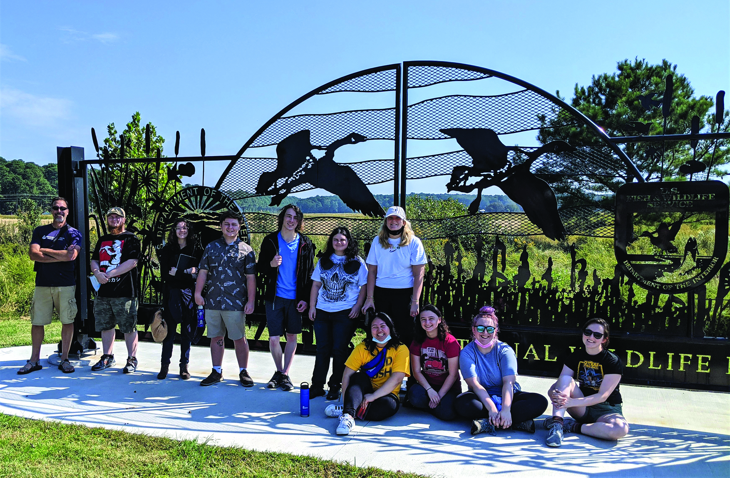 A group of people poses in front of a decorative metal gate featuring bird silhouettes at a wildlife refuge entrance on a sunny day.