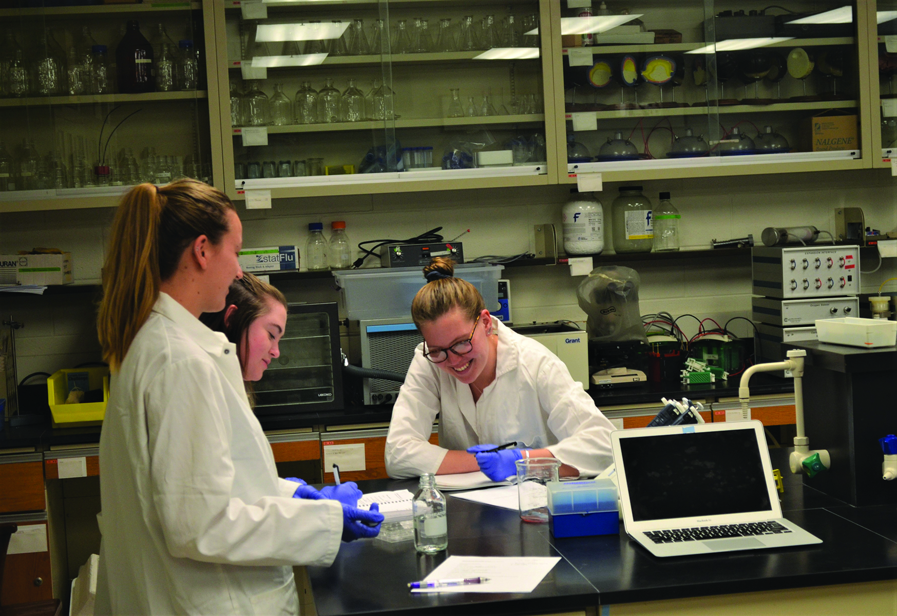 Three women in lab coats and gloves are working in a laboratory. They are engaged in a task involving glass containers and papers.