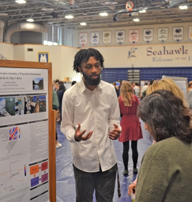 A man in a white shirt discusses a research poster with a woman in a busy gymnasium.