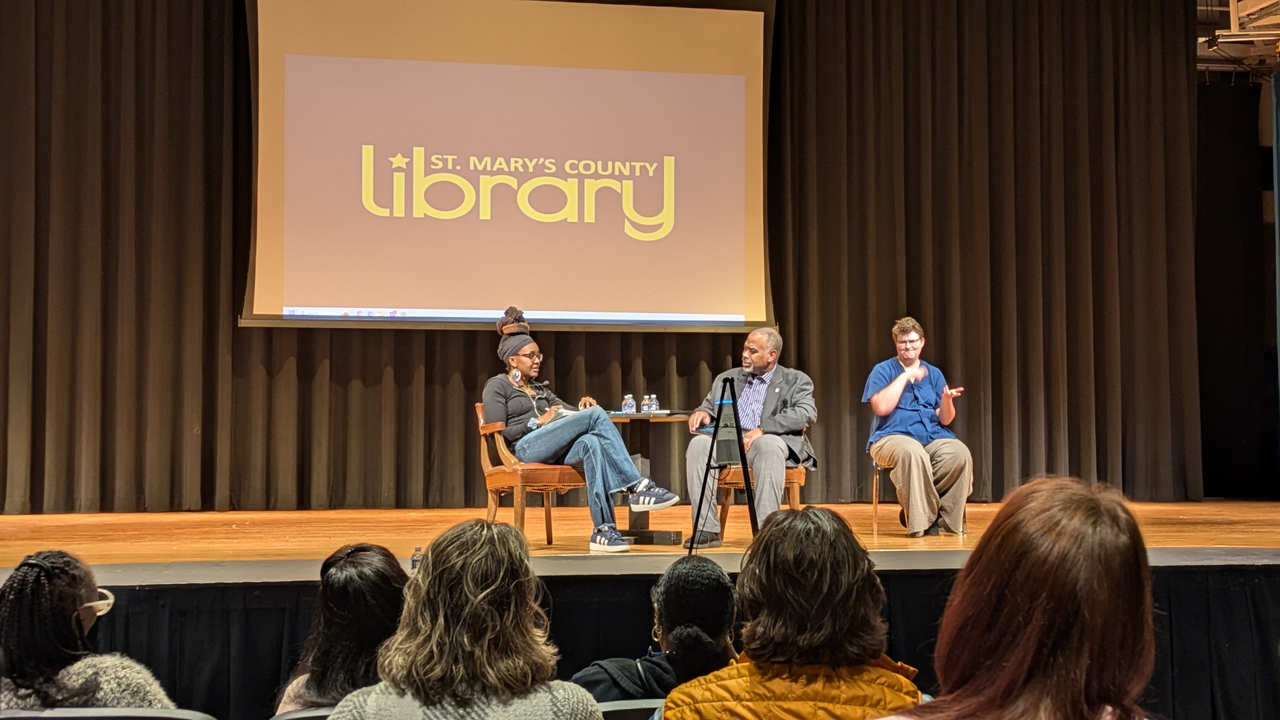 Three people sit onstage at a St. Mary's County Library event. One speaks into a microphone, another listens attentively, 和 third interprets in sign language. The audience is visible in the foreground.