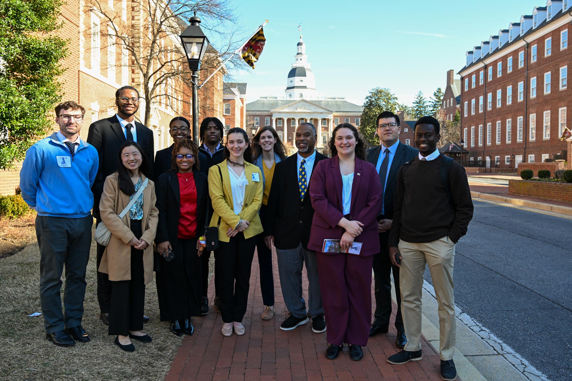 A diverse group of people st和s on a brick pathway in front of a historic building with a dome 和 flags. They are dressed in business 和 casual attire, smiling at the camera, with trees 和 more buildings in the background.