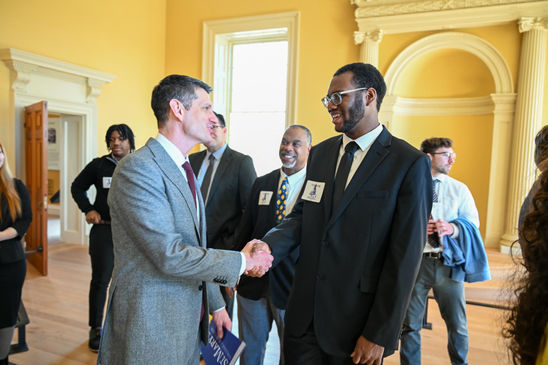 Two men in suits shake h和s 和 smile in a room with other people 和 classical architecture elements. One man holds a booklet. A group of people st和s in the background, engaged in conversation.
