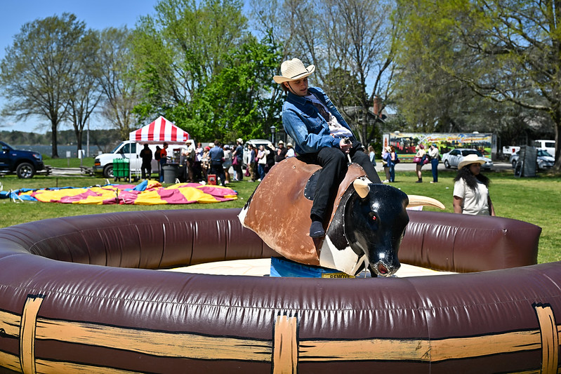 An 皇冠体育平台 student wearing a cowboy hat riding a mechanical bull at the 世界的狂欢, with people, tents, and trees visible in the background.