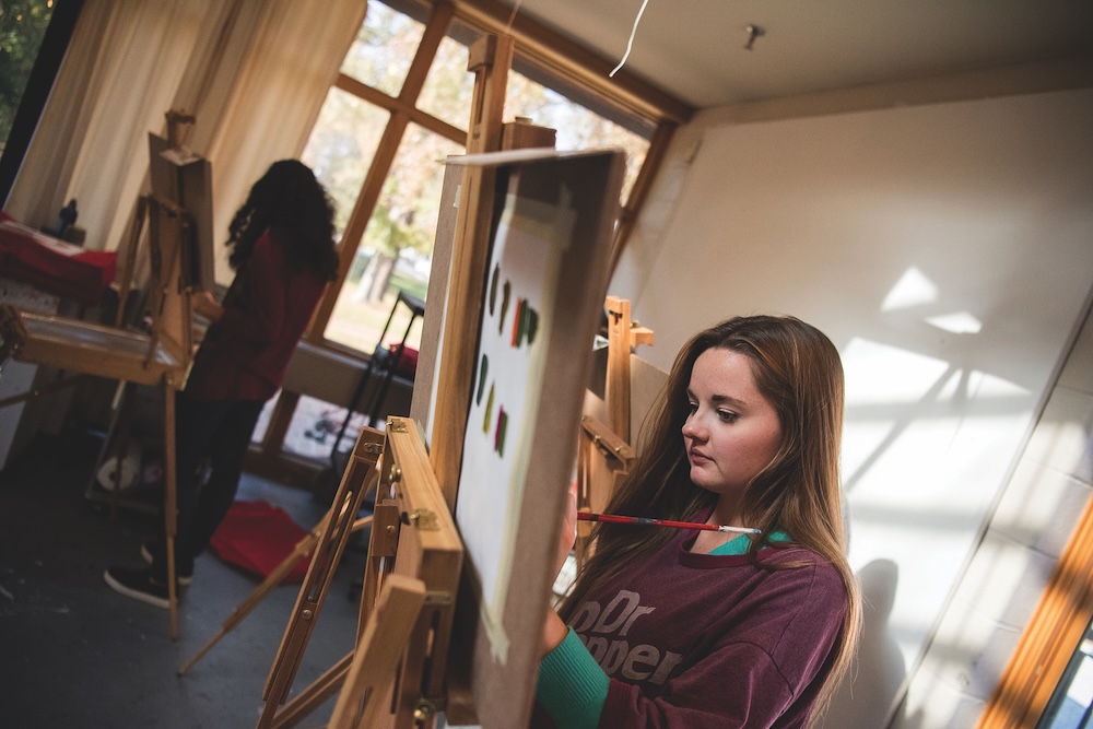 Two people stand at easels painting in a sunlit art studio with large windows. The person in the foreground is focused on their work.