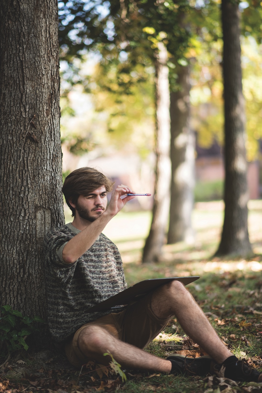 A young man sits on the ground against a tree in a park, holding a pencil and drawing on a sketchpad.