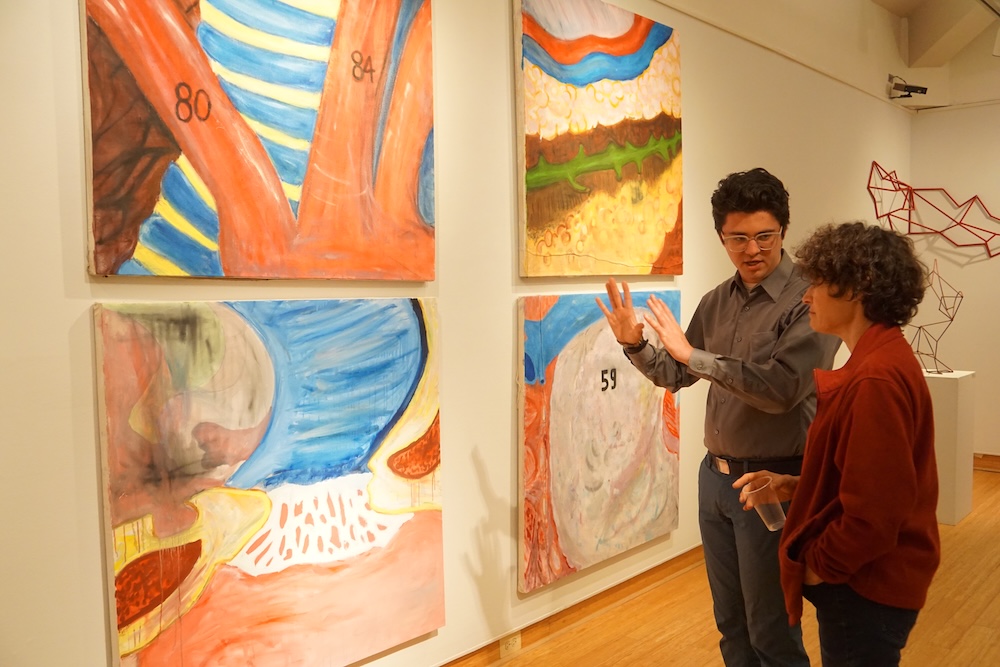 Two people converse in front of colorful abstract paintings displayed on a gallery wall with wooden flooring.