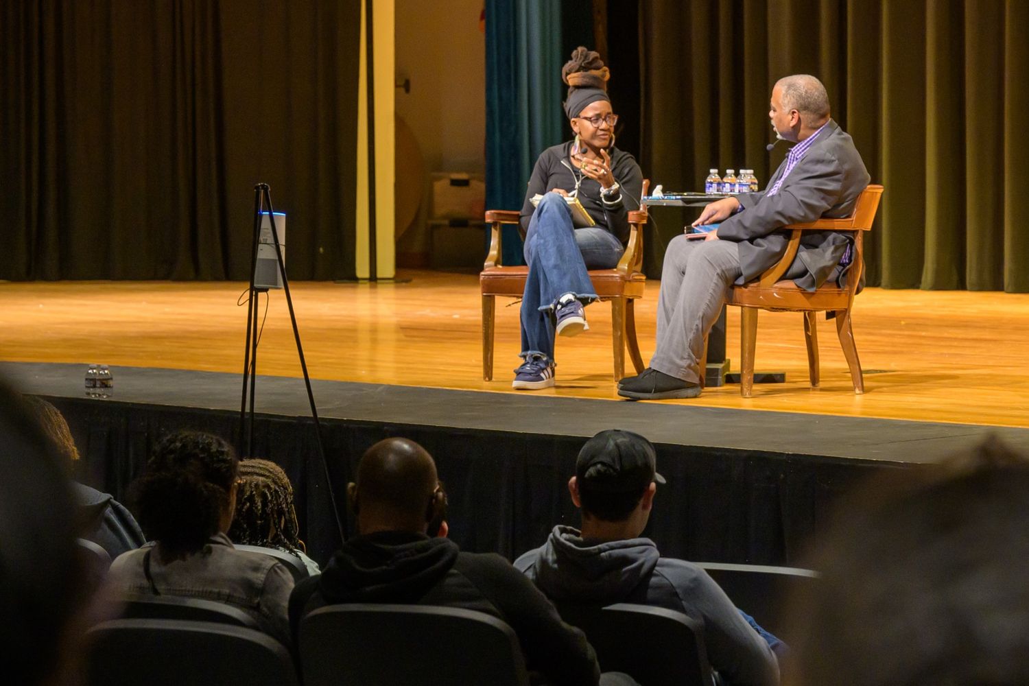 Two people sit on stage having a discussion while an audience watches from the seats in front of them. There are bottled waters on the table between the speakers.
