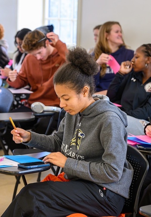 A student writes on a notepad at a desk in a classroom, while other students sit and interact in the background.