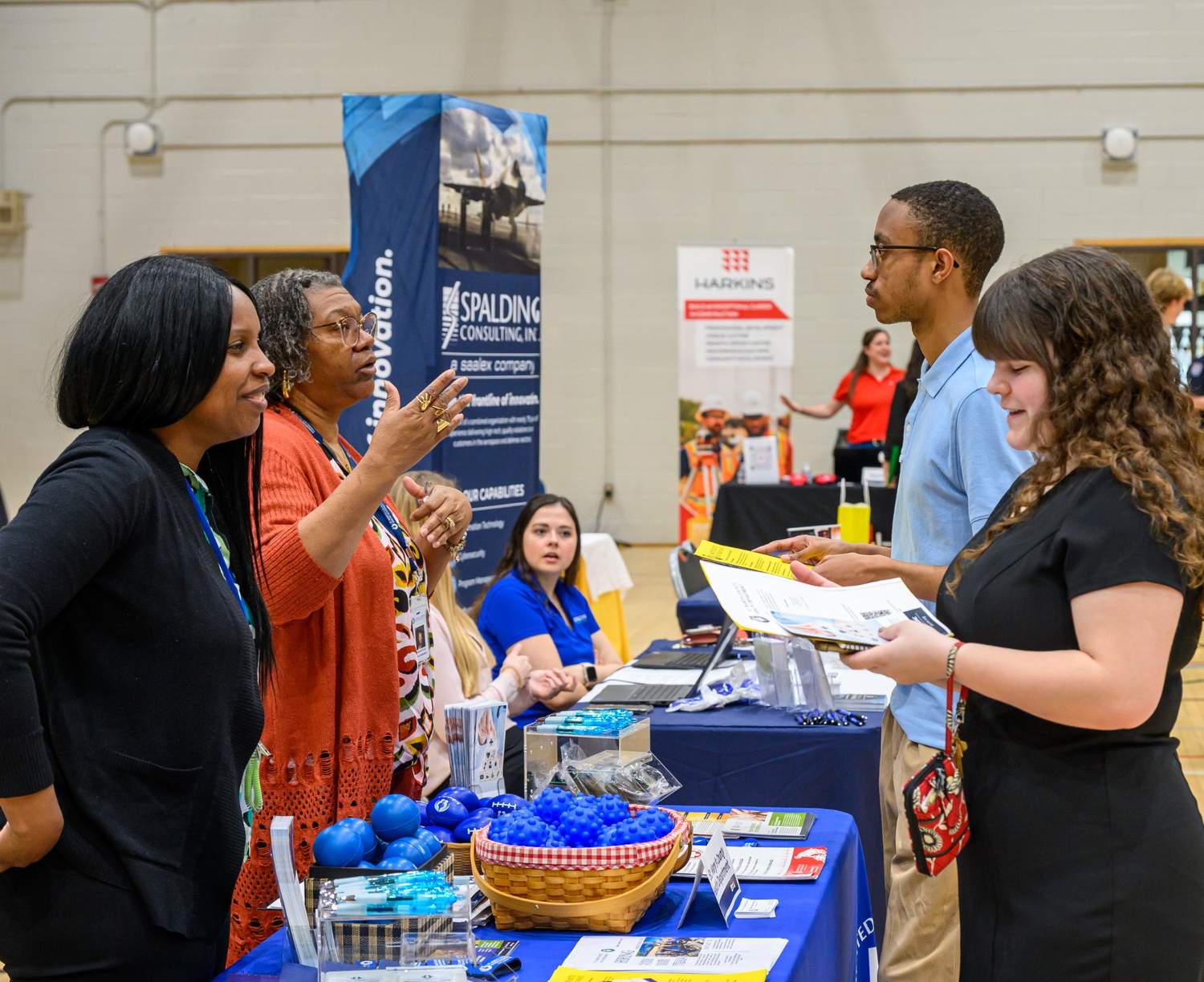 Two women talk to a man and woman at a consulting company booth during an indoor career fair, with promotional materials and brochures displayed on the table.
