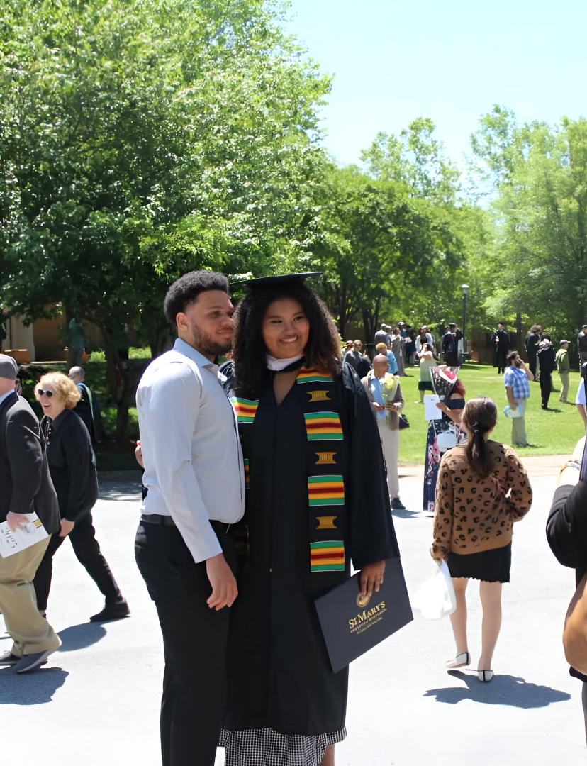 A graduate in cap and gown poses with a man outdoors after a ceremony, surrounded by other people on a sunny day.