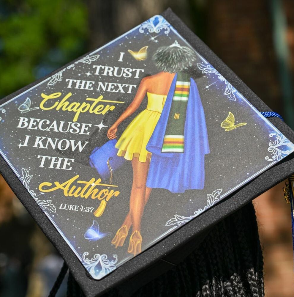 A decorated graduation cap shows an illustration of a woman in a yellow dress with a blue sash and the words, "I trust the next chapter because I know the Author. Luke 1:37.