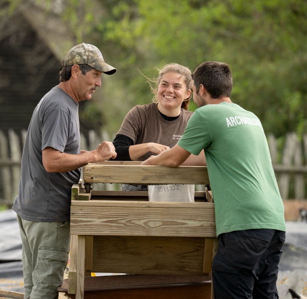 Three people stand around a wooden sifting screen outdoors, working and talking together. Trees and a wooden fence are visible in the background.