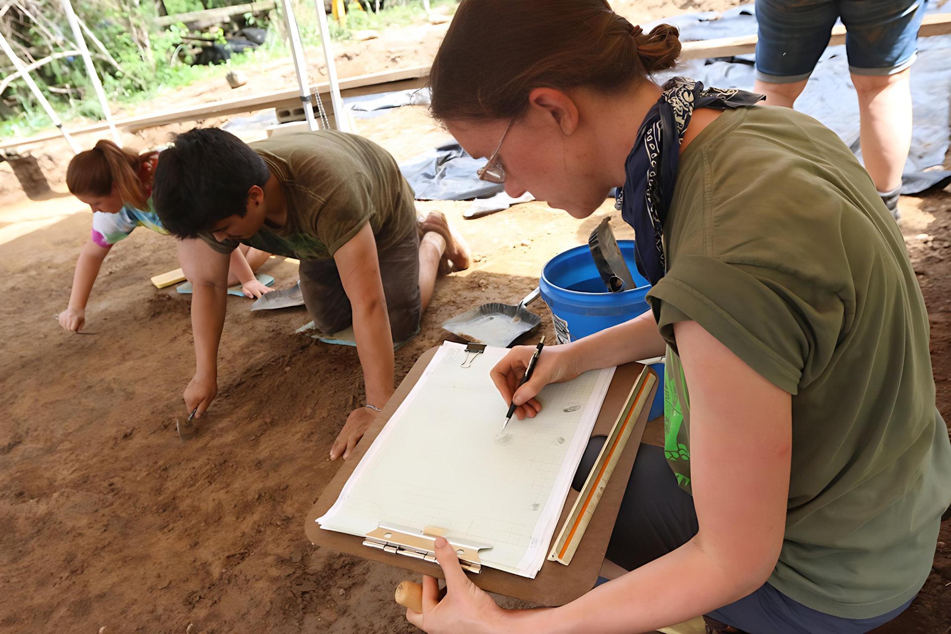 Several people work at an archaeological dig site; one person records findings on a clipboard while others carefully excavate soil in the background.