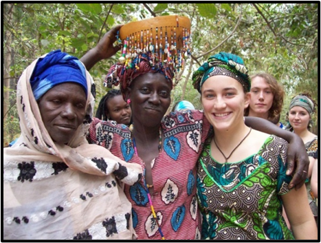Three women pose together outdoors, one holding a decorative item above her head. They are dressed in colorful traditional clothing and headscarves. Other people stand in the background.
